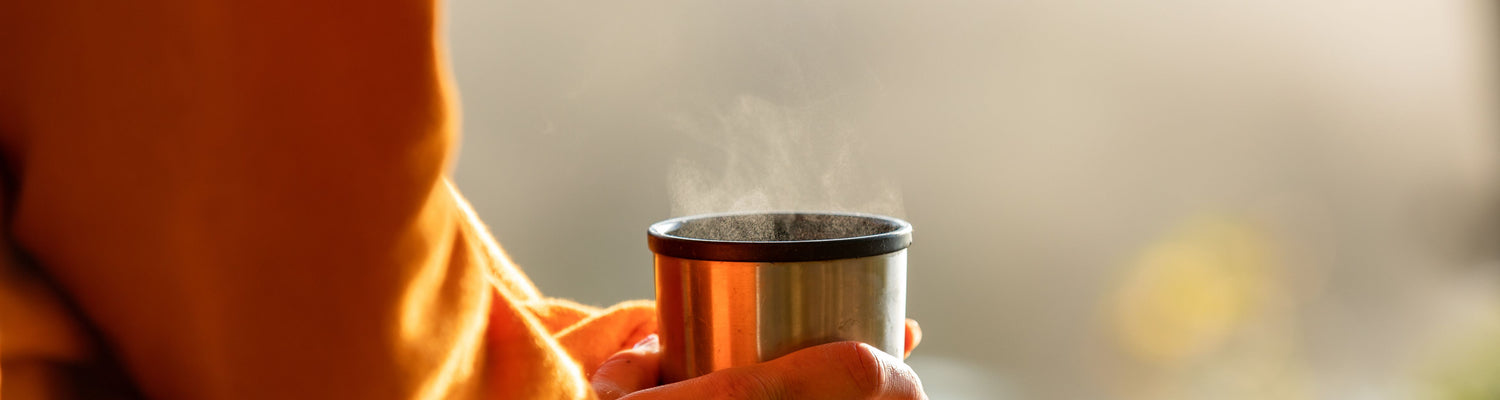 Zu sehen ist ein atmosphärisches Bild mit einer dampfenden Kaffeetasse, die in der Hand gehalten wird